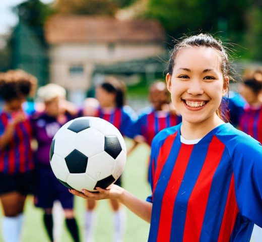 Happy soccer player holding a ball on playing field and looking at  camera while her teammates are in the background.